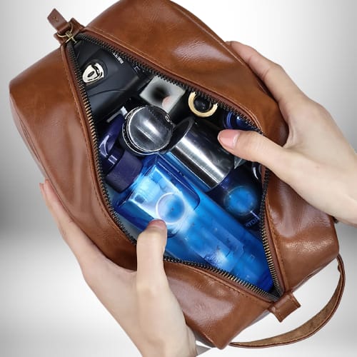 Gros plan d'une personne tenant une trousse de toilette de voyage marron ouverte, qui contient divers articles, notamment une bouteille d'eau bleue.