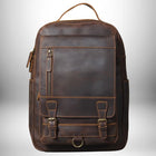 Un sac à dos en cuir marron pour homme est présenté sur un fond légèrement dégradé blanc. Il est orné de coutures apparentes et de détails en métal doré, tels que les fermoirs des poches inférieures et la fermeture éclair de la poche supérieure. Il est pourvu d'une poignée supérieure et de poches latérales.
 — vue 8