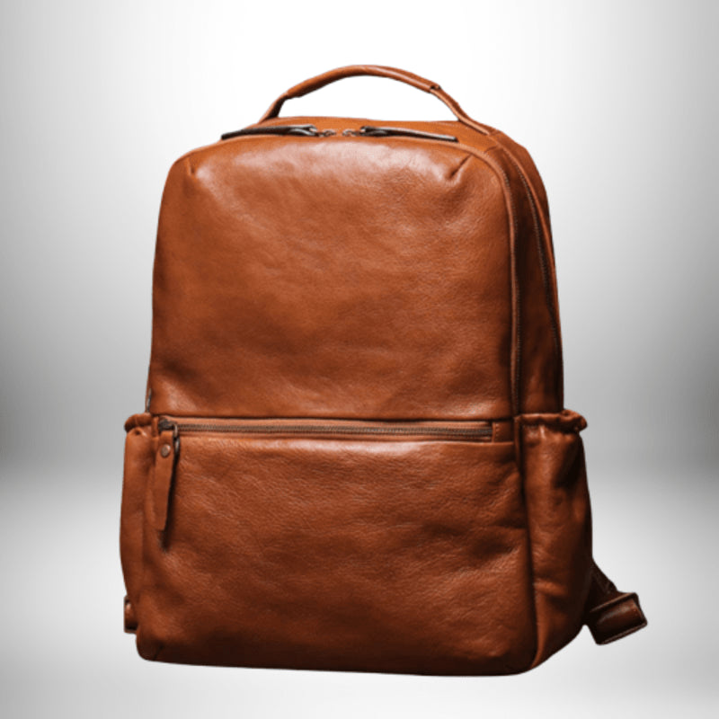 Un sac à dos en cuir marron clair est assis devant un fond dégradé blanc. Il a une poignée sur le dessus et une poche zippée en bas. Les sangles sont repliées derrière le sac à dos.