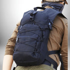 Gros plan d'une personne portant un sac à dos noir. La personne porte un chapeau et une chemise kaki. Le sac à dos a une poche zippée à l'avant, des sangles latérales et des sangles de poitrine. La personne porte également un pantalon vert olive. L'arrière-plan est un mur blanc dégradé. — vue 4