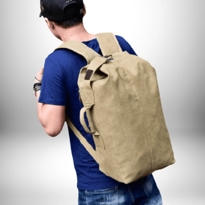 Un homme vu de dos porte un sac à dos beige sur les épaules. Il porte un t-shirt bleu, un chapeau noir et un bracelet. Il se tient devant un fond blanc dégradé.
